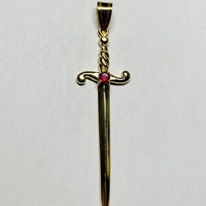 May include: A gold-toned sword pendant featuring a twisted handle design and a small, round, vibrant red gemstone set at the center of the crossguard. The pendant includes a polished bail at the top for attachment to a chain or necklace.