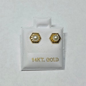 May include: A pair of gold earrings with a pearl in the center. The earrings are in a white box with the text "14KT. GOLD" printed on the bottom.