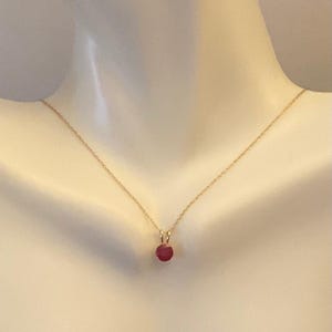 May include: A delicate gold necklace featuring a small, round, ruby pendant. The pendant is a vibrant red color and hangs from a thin gold chain. The necklace is displayed on a mannequin, showcasing its simple elegance.