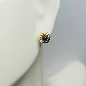 May include: A pair of gold stud earrings with a black gemstone in the center of each earring. The earrings are hexagonal in shape and have a textured design around the gemstone.