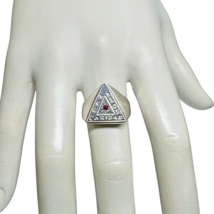 May include: A silver ring with a triangular face. The face has the words "PAZ AMOR CARIDAD" and a small red stone in the center. The ring is on a light-colored hand.