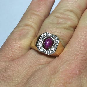 May include: A gold ring with a rectangular face. It features an oval ruby at the centre, encircled by small, clear gemstones. The ring is set on a hand.