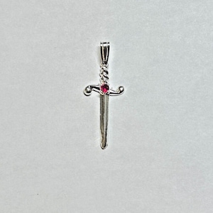 Può includere: Un ciondolo a forma di spada in argento con una pietra preziosa rossa al centro. La spada ha un design attorcigliato e una punta appuntita.