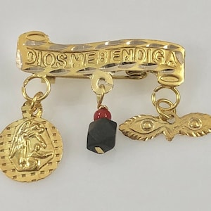 Op de afbeelding: Gouden broche met een decoratieve inscriptie die "DIOS ME BENDIGA" luidt. De broche heeft een centrale hanger met een religieus figuur, een zwarte hanger en een hanger met twee ogen.