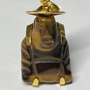 Peut inclure: Pendentif sculpté en œil de tigre représentant une figurine assise portant un chapeau. La figurine a des accents dorés sur le chapeau, les épaules et les pieds. Le pendentif est marron avec des rayures marron foncé.