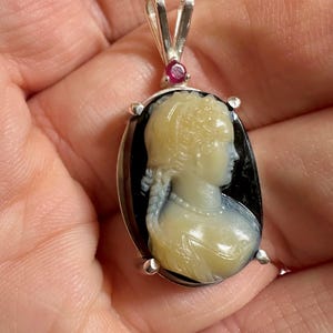 May include: Oval pendant with a cameo of a woman's profile in cream and blue against a black background. The pendant is set in silver with a small red gemstone at the top.