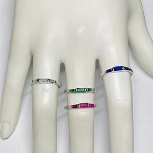 May include: Four silver rings with different colored gemstones. The rings are stacked on a mannequin's finger. The top ring has a blue gemstone, the second ring has a green gemstone, the third ring has a pink gemstone, and the bottom ring has a clear gemstone.