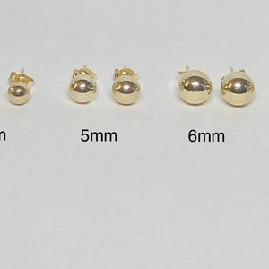May include: A collection of gold ball stud earrings in various sizes: 3mm, 4mm, 5mm, 6mm, and 8mm. The earrings are displayed on a white surface, showcasing their polished, reflective finish. The sizes are labeled below each pair.