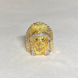 May include: A gold ring with a detailed design of an Native American chief wearing a headdress. The chief's face is highlighted with blue and red gemstones.
