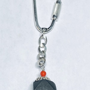 May include: A silver keychain with a black, geometric charm and a red bead. The charm has a silver ring around the top.