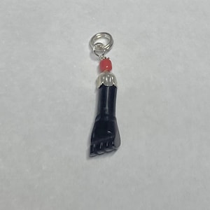 Puede incluir: Un dije de pie negro con una tapa plateada y una cuenta roja, suspendido de un anillo plateado. El dije está detallado con dedos y una superficie texturizada.