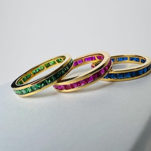 Puede incluir: Tres anillos de oro con una banda de piedras preciosas de colores. Los anillos están apilados uno encima del otro. El anillo superior tiene piedras azules, el anillo del medio tiene piedras rosas y el anillo inferior tiene piedras verdes.