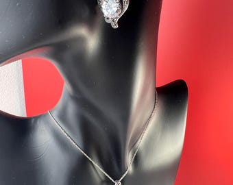 Peut inclure: Un ensemble de bijoux argentés comprenant un collier pendentif en forme de cœur et des boucles d'oreilles assorties. Chaque pièce comporte une pierre précieuse centrale et transparente entourée d'un motif ajouré décoratif. L'ensemble est présenté sur un mannequin noir.