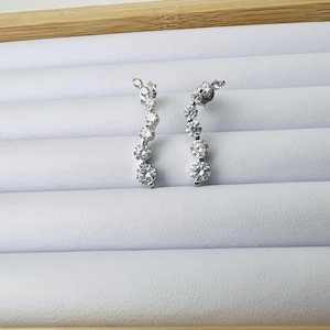 May include: A pair of silver-toned drop earrings. Each earring features a series of round, clear gemstones graduating in size. The earrings are displayed on a white surface within a wooden frame.
