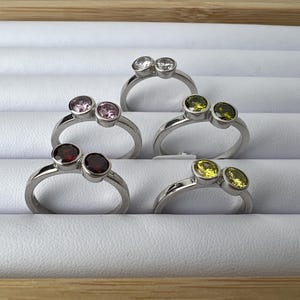 May include: Five silver rings with double gemstones. The rings feature clear, pink, red, yellow, and green gemstones.