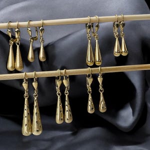 May include: Six pairs of gold dangle earrings with teardrop-shaped pendants. The earrings are displayed on a wooden stand.