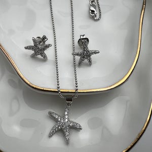 May include: A silver necklace and earrings set featuring a starfish pendant and matching earrings. The starfish is adorned with sparkling clear stones.