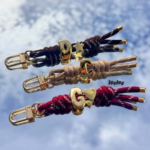 May include: Three personalized keychains with gold-colored hardware and initial charms. The keychains feature braided cords in brown, tan, and burgundy, each with a different initial and charm: "D" with a star, "C", and "G" with a heart. The word "leelee" is in the image.