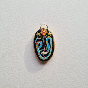 May include: A black oval pendant with a gold rim and a colorful design. The design features a flower with a pink center and orange petals, a blue and green snake-like figure, and two gold stars.