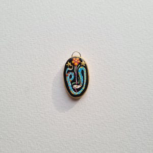 May include: A black oval pendant with a gold rim and a colorful design of a snake and a flower. The snake is blue and yellow, and the flower is orange and pink.