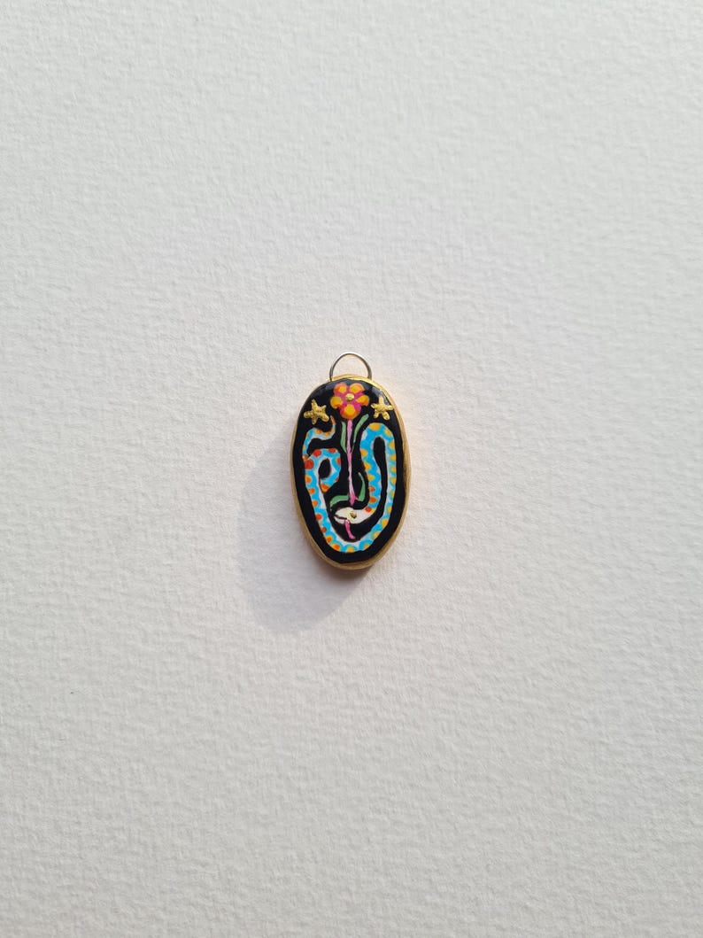 May include: A black oval pendant with a gold rim and a colorful design of a snake and a flower. The snake is blue and yellow, and the flower is orange and pink.