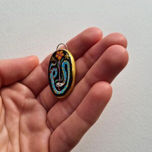 May include: A gold-painted oval pendant with a black background and a colorful design featuring a blue snake, a red flower, and gold stars.