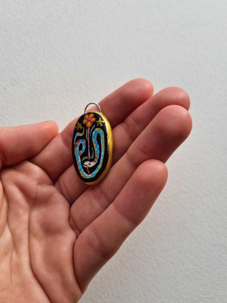 May include: A gold-painted oval pendant with a black background and a colorful design featuring a blue snake, a red flower, and gold stars.