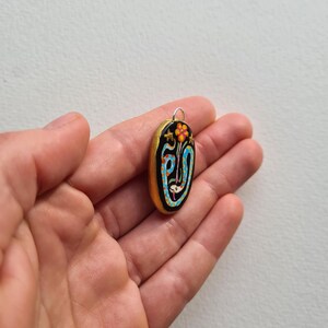 May include: A gold oval pendant with a black background and a colorful design featuring a flower, stars, and a blue and white snake.