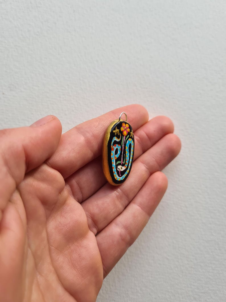 May include: A gold oval pendant with a black background and a colorful design featuring a flower, stars, and a blue and white snake.