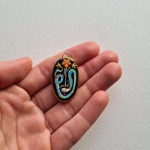 May include: A black oval pendant with a gold rim, featuring a colorful painted design of a flower with a blue and white snake wrapped around it. The pendant has a gold loop at the top for attaching a chain.
