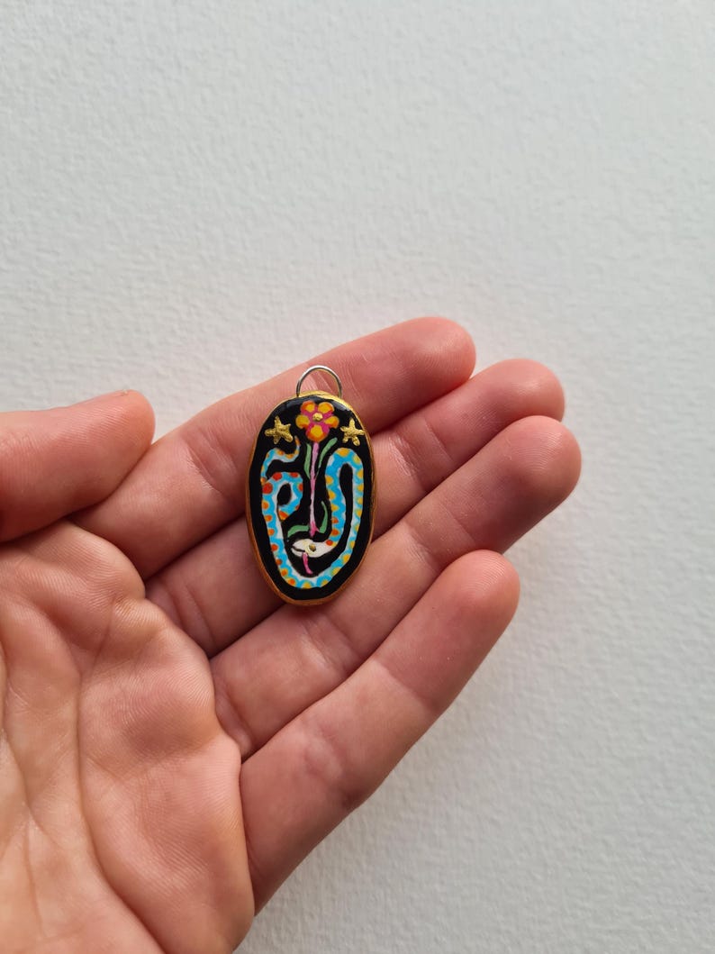 May include: A black oval pendant with a gold rim, featuring a colorful painted design of a flower with a blue and white snake wrapped around it. The pendant has a gold loop at the top for attaching a chain.