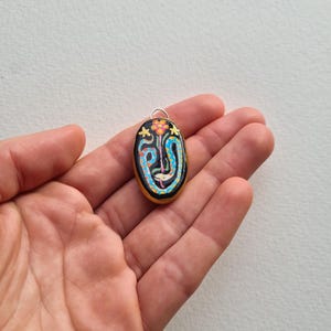May include: A black oval pendant with a colorful snake design and a small flower on top. The pendant is made of polymer clay and has a silver bail.