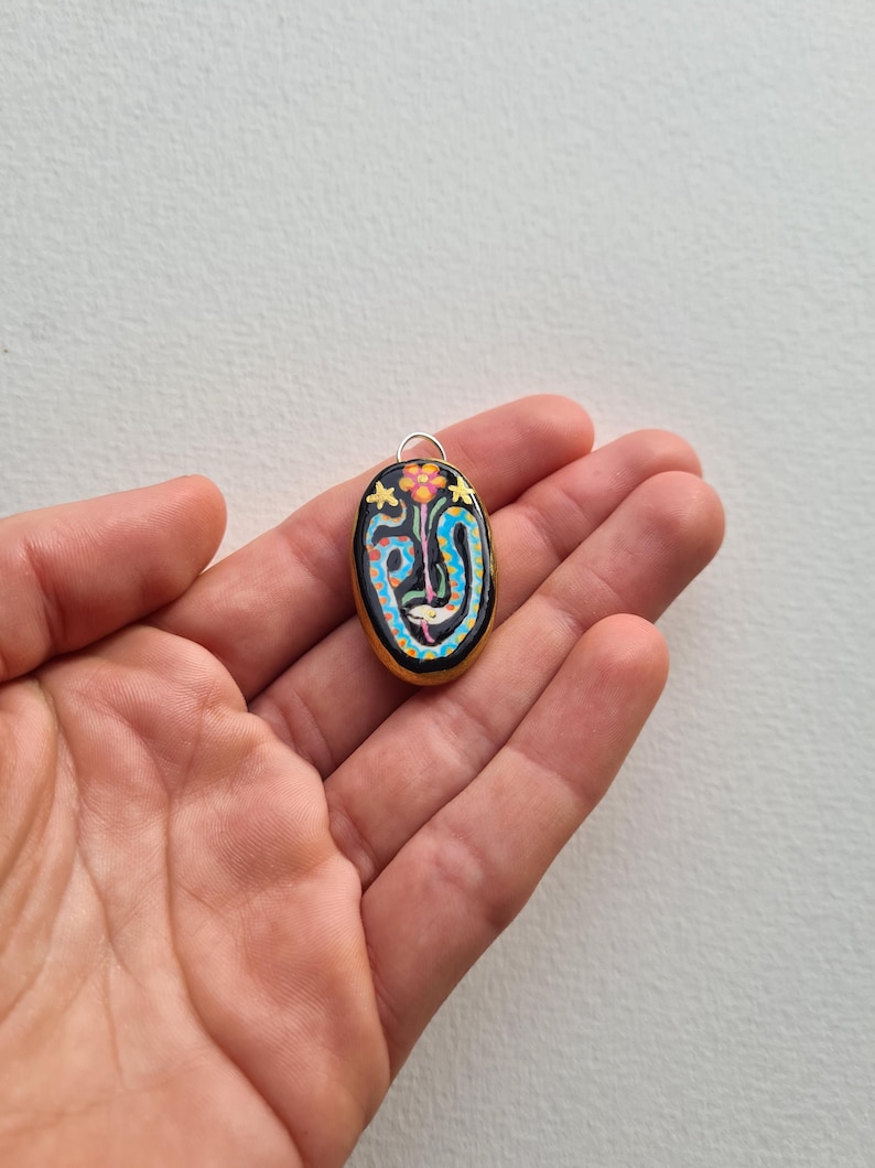 May include: A black oval pendant with a colorful snake design and a small flower on top. The pendant is made of polymer clay and has a silver bail.