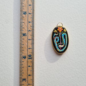 May include: A gold-toned pendant with a black oval-shaped charm featuring a colorful snake design with a flower and stars. The snake is blue, white, and orange, and the flower is orange and yellow. The stars are gold.