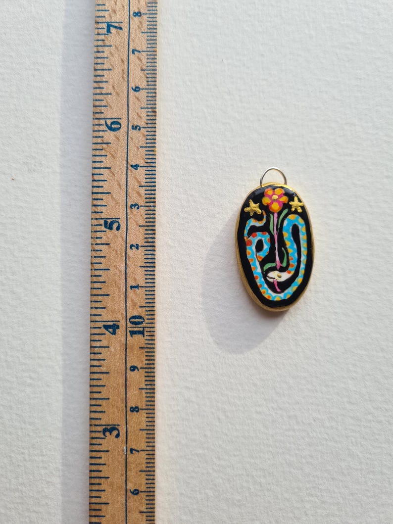 May include: A gold-toned pendant with a black oval-shaped charm featuring a colorful snake design with a flower and stars. The snake is blue, white, and orange, and the flower is orange and yellow. The stars are gold.