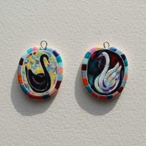 May include: Two oval pendants with painted swan designs. One pendant features a black swan against a colorful background, the other a white swan. Both have a colorful border and a silver loop for hanging.