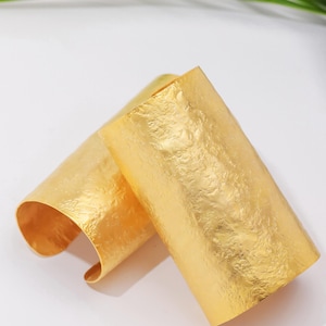 May include: Two gold-coloured, textured metal cuff bracelets. The bracelets are rectangular with a rolled edge, and are displayed on a white surface. The bracelets have a hammered metal texture.