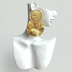 May include: Gold-coloured statement earrings with a textured, hammered metal finish. The earrings feature a large, circular pendant and a smaller, diamond-studded circle above. The earrings are displayed on a white mannequin head.