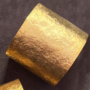 May include: A wide, textured gold cuff bracelet. The surface of the bracelet has a hammered or crumpled appearance, reflecting light. The jewellery piece is a bold statement accessory.