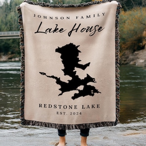 May include: A tan woven throw blanket with fringe. The blanket features a black silhouette of a lake with the text "Johnson Family Lake House" and "Redstone Lake Est. 2024".