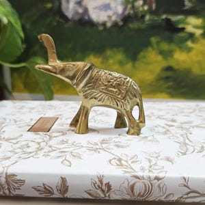 May include: A small, gold-coloured brass elephant figurine with detailed carvings on its back. The elephant is standing on a stack of books with floral patterned covers. The background includes a painting of a landscape and some greenery.