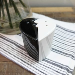 Peut inclure: Un ensemble de salière et poivrière en forme de cœur. Un côté est noir, l'autre est blanc. L'ensemble est posé sur un tissu à rayures blanches et grises. La salière a deux trous et la poivrière en a un.