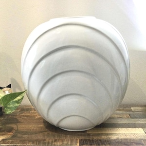 Peut inclure: Un grand vase rond en céramique blanche avec un motif en forme de coquillage. Le vase présente une série de crêtes incurvées concentriques qui créent une surface texturée. Le vase est posé sur une surface en bois.