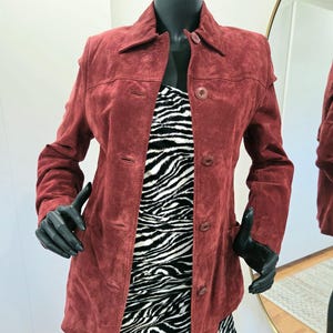 May include: A dark red suede jacket featuring a classic collar, long sleeves, and a front button fastening. The jacket is worn over a black and white zebra-patterned dress. The suede fabric has a matte texture, and the buttons are round and matching.