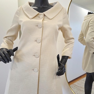 May include: A cream-coloured coat with a rounded collar and large, matching buttons. The coat is displayed on a mannequin with black gloves. A mirror in the background reflects the coat's back. The coat has long sleeves and a tailored fit.