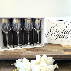 May include: A set of four clear glass wine glasses with stems, each featuring a gold circular sticker with the text Cristal d'Arques France. The glasses are housed in a white box with the brand name Cristal d'Arques printed in black script.