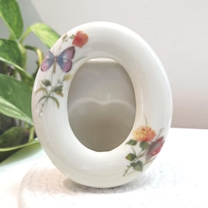 May include: Oval-shaped white ceramic picture frame with floral and butterfly designs. The frame has a smooth, glossy finish and features a central oval opening. The floral and butterfly designs are in shades of red, yellow, purple, and green.