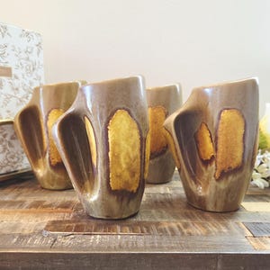 May include: A set of four ceramic mugs featuring a tapered, angular silhouette with an integrated handle. The mugs have a glossy, multi-toned glaze in shades of earthy brown and mustard yellow, with a textured, mottled finish on the exterior surface.