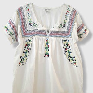 May include: White embroidered top with a V-neck and short sleeves. The top features colourful floral embroidery along the neckline, sleeves, and bodice. The embroidery includes red, blue, yellow, and green thread. The top is made of a lightweight fabric.