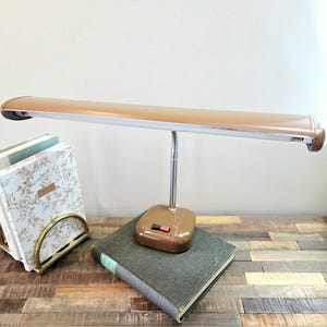 May include: A brown desk lamp with a long, horizontal light head and a flexible silver neck. The base is square and brown, featuring a rectangular black and red power switch. The lamp has a smooth, metallic finish and a simple, functional design.